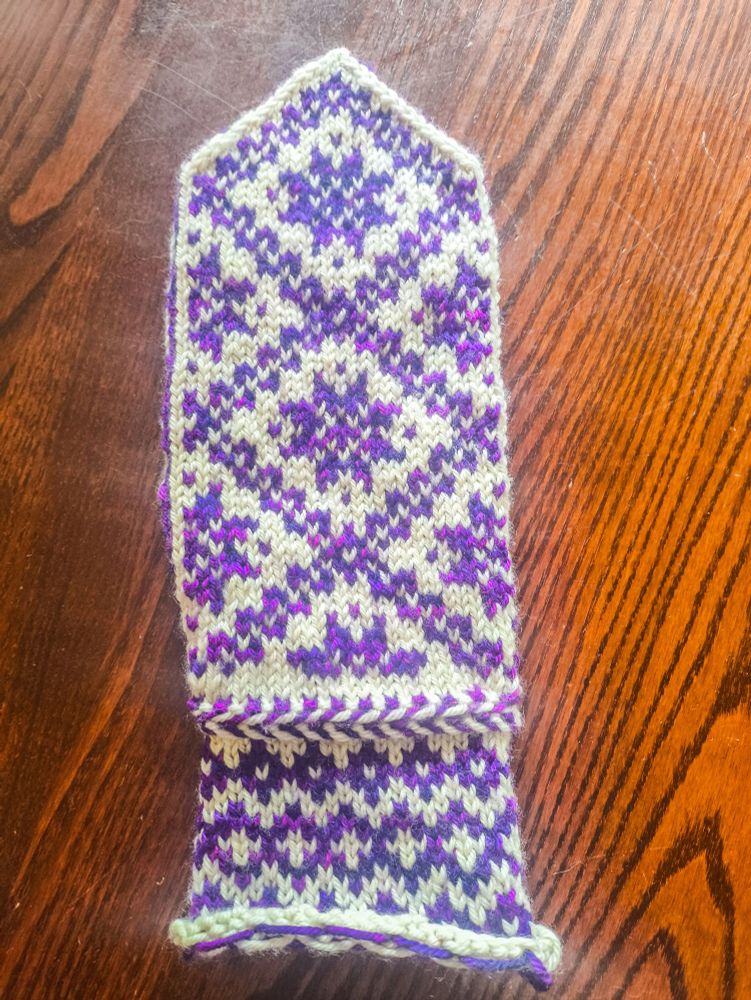The completed front of a partially-knitted mitten knit in white wool as the base colour and variegated purple-magenta wool as the contrast colour. The pattern on the wrist is three rows of purple diamonds, with a slightly larger middle row that has white crosses in the middle of the diamonds. Above the wrist is a Latvian braid, a knitting technique that produces a raised, horizontal stripe in alternating colours (it looks similar to a fishtail hair braid). The main body of the mitten is made up of several large diagonally-oriented squares pieced together with eight-pointed stars or snowflakes (I'm not quite sure which they're supposed to be) inside.