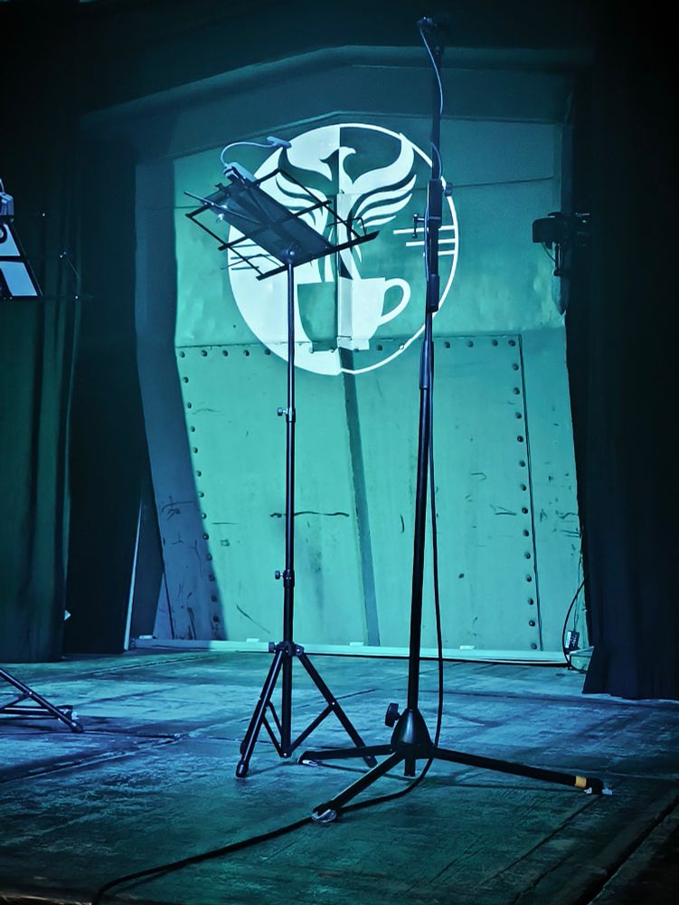 A close up shot of a darkened stage with a metal background. A pale green light shines against the back of the stage. The Amelia Project logo, a half-black, half-white drawing of a phoenix emerging from a coffee cup is also projected on the back of the stage. On the stage is a microphone stand and a stand to hold the script.