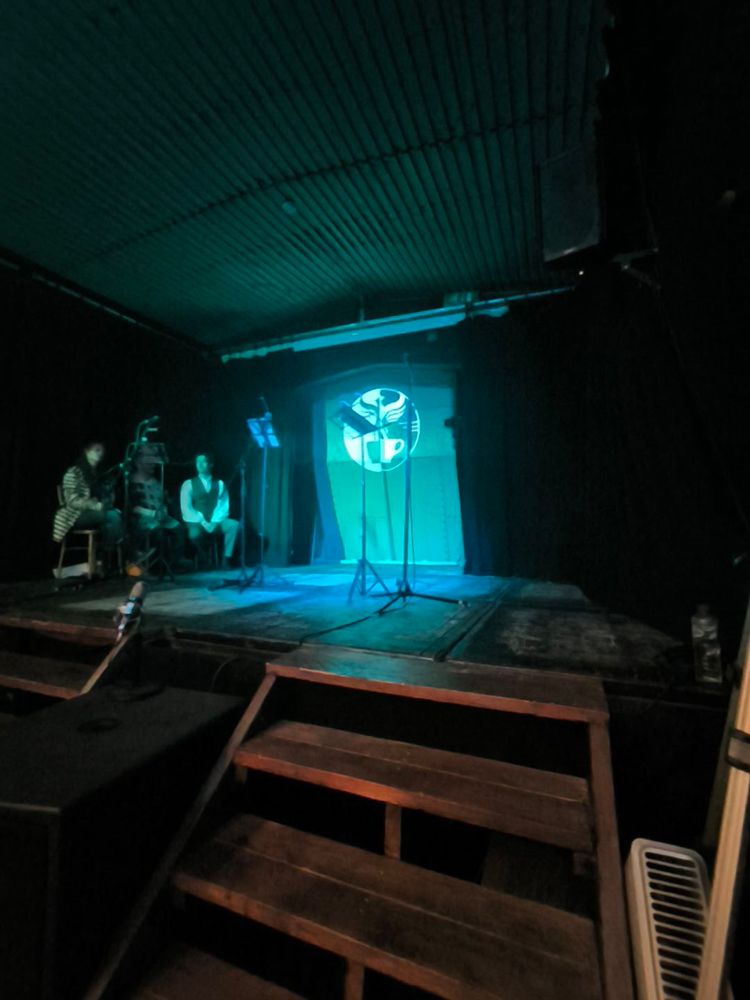 A distance shot of a darkened stage with a metal background. A pale green light shines against the back of the stage. The Amelia Project logo, a half-black, half-white drawing of a phoenix emerging from a coffee cup is also projected on the back of the stage. On the stage is a microphone stand and a stand to hold the script. On the left edge of the stage three people, the main cast of the show, are sitting.