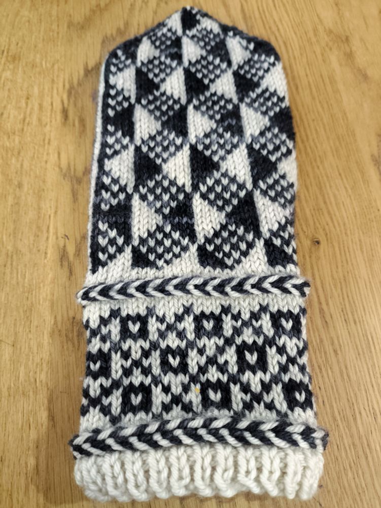 An almost complete mitten-in-progress knit in white wool as the base colour and semi-solid black/charcoal grey wool as the contrast colour. The entirety of the body is knit, leaving only the thumb. 

The wrist from the bottom up: a small section of 1x1 rib, then a Latvian braid (a knitting technique that carries the yarn in front and twists it around itself to create horizontal "braids" in your knitting), a colourwork section made up of squares with white centres in the middle of diamonds created by a large crosshatch pattern, then another Latvian braid.

The body from the wrist up: a geometric pattern made up of solid black and white alternating vertical half-diamonds with diamonds between them made up of alternating black and white stitches.
