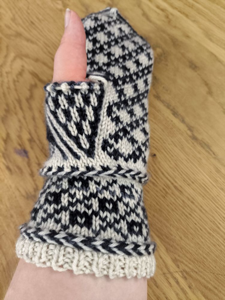 An almost complete mitten-in-progress knit in white wool as the base colour and semi-solid black/charcoal grey wool as the contrast colour. The entirety of the body is knit, leaving only the thumb. It's being tried on by a person with a pale arm holding their hand out so a photo can be taken. Their thumb is sticking out through the hole where the mitten's thumb will later be.

The wrist from the bottom up: a small section of 1x1 rib, then a Latvian braid (a knitting technique that carries the yarn in front and twists it around itself to create horizontal "braids" in your knitting), a colourwork section made up of squares with white centres in the middle of diamonds created by a large crosshatch pattern, then another Latvian braid.

The body from the wrist up: a striped black and white triangular thumb gusset on the left of the palm (to fit the left hand), next to a pattern of white diamonds formed by crossed lines of small black pluses. The thumb gusset currently ends with a hole in the work as the thumb will be knit later. Above the thumb gusset the pattern of diamonds and pluses continues across the whole palm.