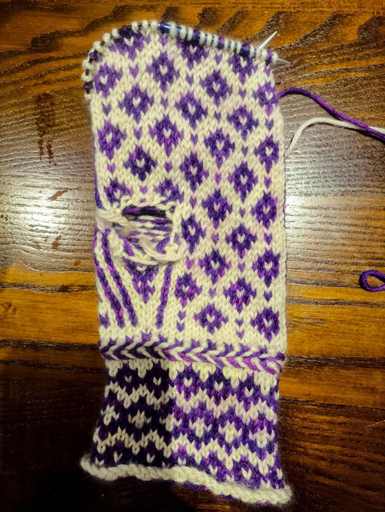 Half of a finished mitten, knit in white yarn as the base colour and bright purple yarn with magenta flashes as the contrast colour, facing palm up on a table. It has a repeating pattern of diamonds with a white stitch in the centre. Some pink splotches, which are dye from the purple yarn that has run because the project got wet, are faintly visible. 