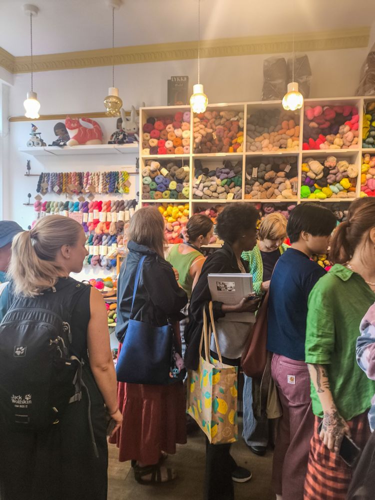 The inside of Loop London, a yarn shop in Islington. Part of a modular cube shelf filled with hanks of yarn of all colours and some small shelves next to it with mini skeins hanging from it and some baskets underneath are visible in the background.  The shop is filled with people waiting to pay. 