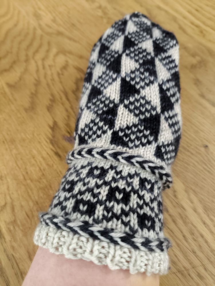 An almost complete mitten-in-progress knit in white wool as the base colour and semi-solid black/charcoal grey wool as the contrast colour. The entirety of the body is knit, leaving only the thumb. It's being tried on by a person with a pale arm holding their hand out so a photo can be taken.

The wrist from the bottom up: a small section of 1x1 rib, then a Latvian braid (a knitting technique that carries the yarn in front and twists it around itself to create horizontal "braids" in your knitting), a colourwork section made up of squares with white centres in the middle of diamonds created by a large crosshatch pattern, then another Latvian braid.

The body from the wrist up: a geometric pattern made up of solid black and white alternating vertical half-diamonds with diamonds between them made up of alternating black and white stitches.