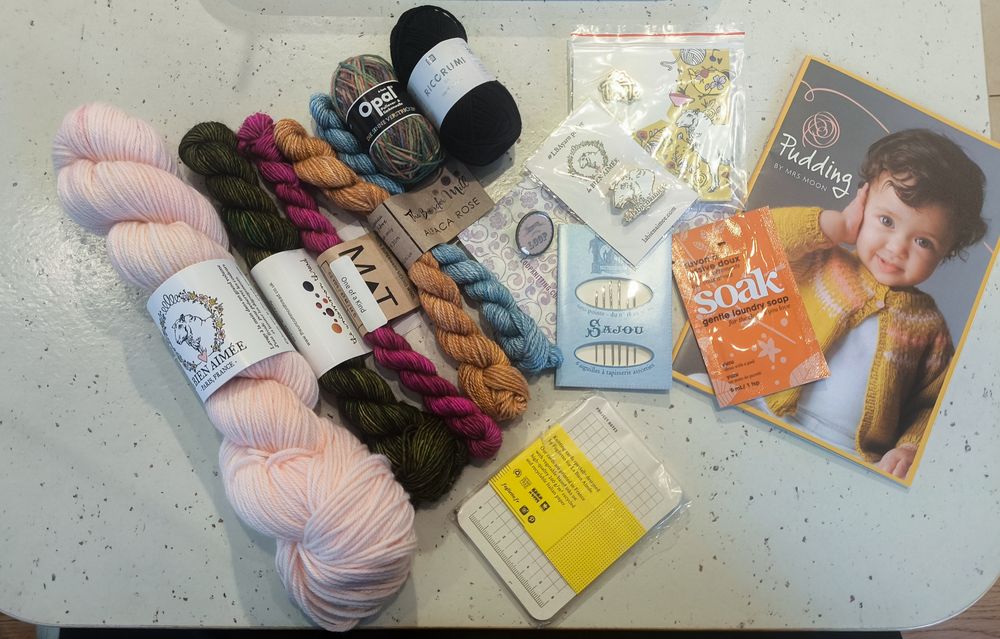 A selection of yarn, notions, and other goodies laid out on a white table with brown speckles. The leftmost skein is a full sized skein in pastel pink from la Bien Aimée. Next to that is a selection of mini skeins of various sizes: a deep forest green one from The Uncommon Thread (colourway: undergrowth), then a deep magenta one from Madelinetosh (colourway: one of a kind (?)), an Alpaca Rose set from The Border Mill (colourway name unknown, dyed specially for the Loop 20th anniversary), a variegated mini ball from Opal, and a black mini ball of Ricorumi. Next to the mini skeins are a selection of notions and other little goodies: a Loop London badge, a la Bien Aimée badge featuring the sheep head from their logo, another la Bien Aimée badge with some text (glare from the lighting has obscured it), a packet of darning needles, a small sachet of Soak gentle laundry soap in the yuzu scent, a pack of knitting note cards designed for la Bien Aimée, and a pattern booklet for (I believe) some children's knits. The booklet is called Plump by Mrs Moon (a yarn brand) and features a toddler wearing a mustard yellow cardigan with some peach and white details.