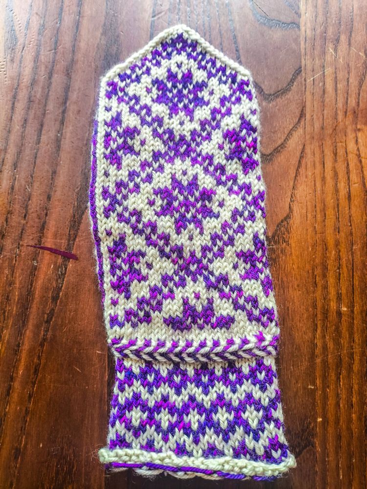 The completed front of a mitten knit in white wool as the base colour and variegated purple-magenta wool as the contrast colour. The pattern on the wrist is three rows of purple diamonds, with a slightly larger middle row that has white crosses in the middle of the diamonds. Above the wrist is a Latvian braid, a knitting technique that produces a raised, horizontal stripe in alternating colours (it looks similar to a fishtail hair braid). The main body of the mitten is made up of several large diagonally-oriented squares pieced together with eight-pointed stars or snowflakes (I'm not quite sure which they're supposed to be) inside.