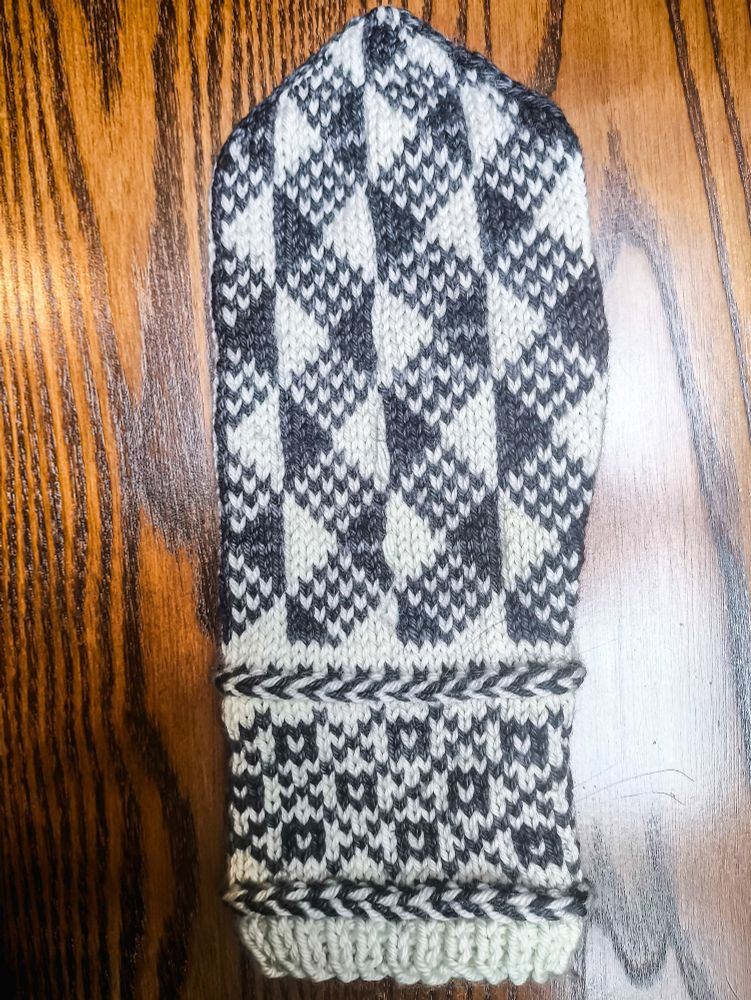 A complete mitten-in-progress knit in white wool as the base colour and semi-solid black/charcoal grey wool as the contrast colour.

The wrist from the bottom up: a small section of 1x1 rib, then a Latvian braid (a knitting technique that carries the yarn in front and twists it around itself to create horizontal "braids" in your knitting), a colourwork section made up of squares with white centres in the middle of diamonds created by a large crosshatch pattern, then another Latvian braid.

The body from the wrist up: a geometric pattern made up of solid black and white alternating vertical half-diamonds with diamonds between them made up of alternating black and white stitches.