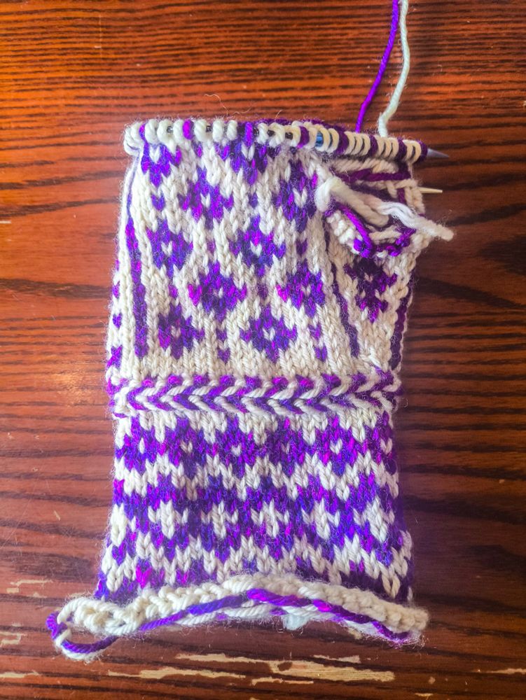 The back of a partially-knitted mitten knit in white wool as the base colour and variegated purple-magenta wool as the contrast colour. The pattern on the wrist is three rows of purple diamonds, with a slightly larger middle row that has white crosses in the middle of the diamonds. Above the wrist is a Latvian braid, a knitting technique that produces a raised, horizontal stripe in alternating colours (it looks similar to a fishtail hair braid). The main body of the mitten is made up of a repeating pattern of diamonds with white centres with single contrast-colour stitches between them.
