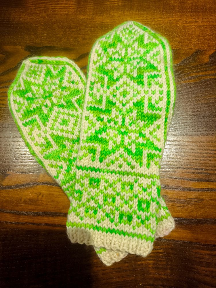 A finished pair of mittens knit in white and bright, neon highlighter green yarn with two large, eight-pointed stars on each hand and a crosshatched pattern on the wrists. The right mitten rests on top of the left at an angle.