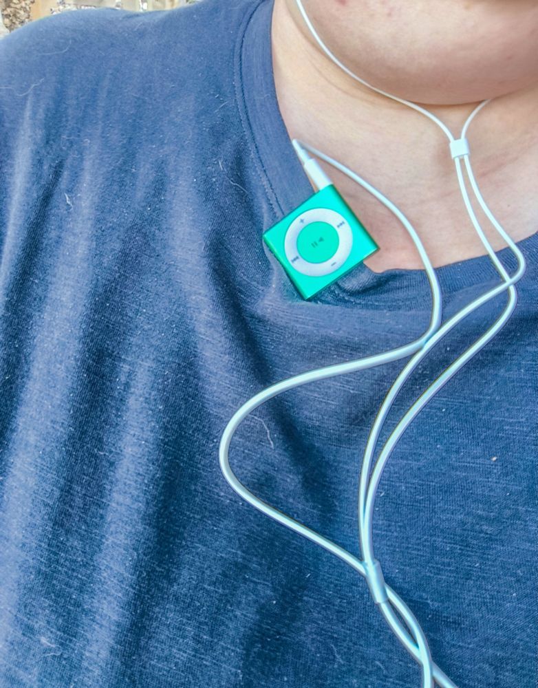 A pale person in a stonewashed(?) style black crew neck t-shirt. The shot is a close up so all that can be seen is their neck and t-shirt, with a bright green Apple iPod 4th generation Shuffle (a small, square MP3 player that clips onto your shirt or similar) clipped to the neckline with an earphone cable.