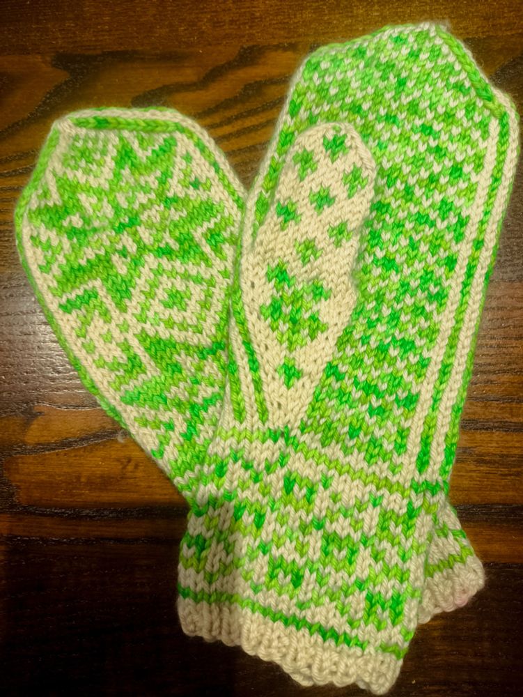 A finished pair of mittens knit in white and bright, neon highlighter green yarn. The bottom mitten is facing upwards with two large, eight-pointed stars on the hand and a crosshatched pattern on the wrist. The top mitten is facing palm-up, showing a pattern of small repeating crosses across the palm. The left mitten rests on top of the right at an angle.