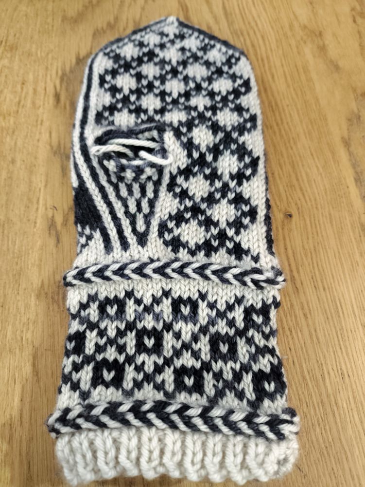An almost complete mitten-in-progress knit in white wool as the base colour and semi-solid black/charcoal grey wool as the contrast colour. The entirety of the body is knit, leaving only the thumb. 

The wrist from the bottom up: a small section of 1x1 rib, then a Latvian braid (a knitting technique that carries the yarn in front and twists it around itself to create horizontal "braids" in your knitting), a colourwork section made up of squares with white centres in the middle of diamonds created by a large crosshatch pattern, then another Latvian braid.

The body from the wrist up: a striped black and white triangular thumb gusset on the left of the palm (to fit the left hand), next to a pattern of white diamonds formed by crossed lines of small black pluses. The thumb gusset currently ends with a hole in the work as the thumb will be knit later. Above the thumb gusset the pattern of diamonds and pluses continues across the whole palm.
