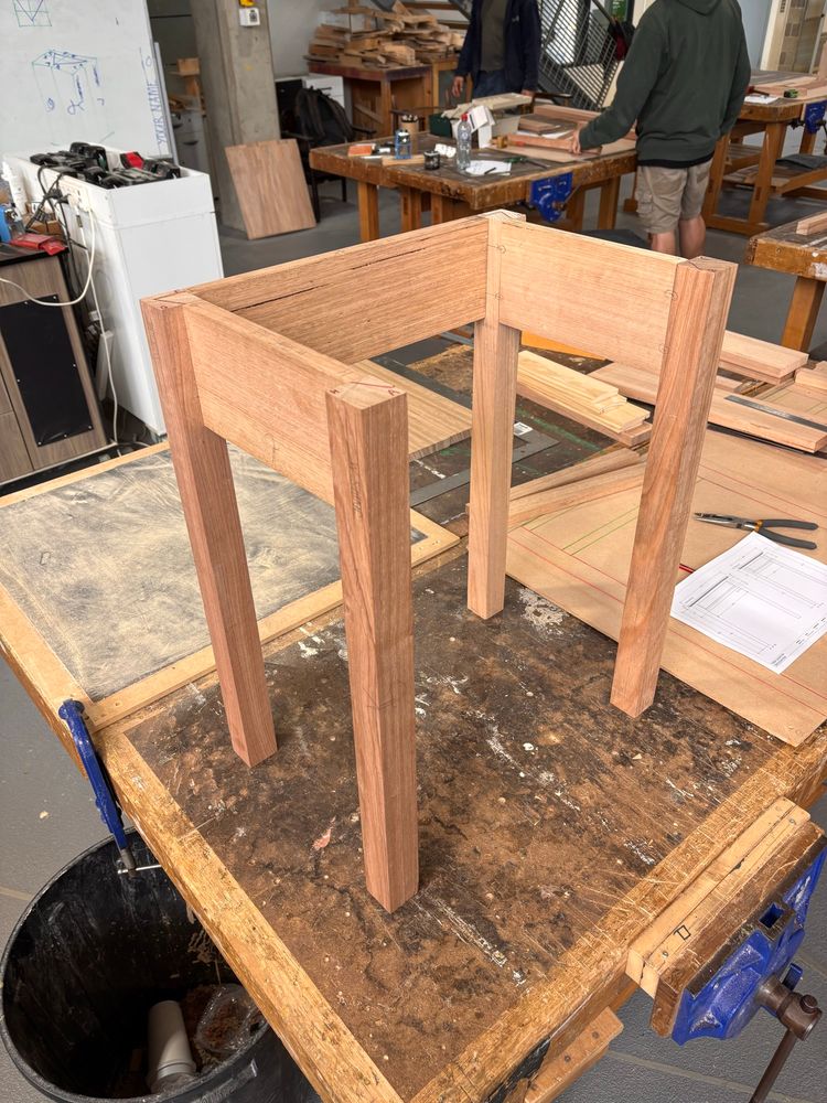 A work in progress shot of a a half built bedside table. The two sides and back are assembled and upright, with the front cross rails missing. It stands on a timber workbench. 