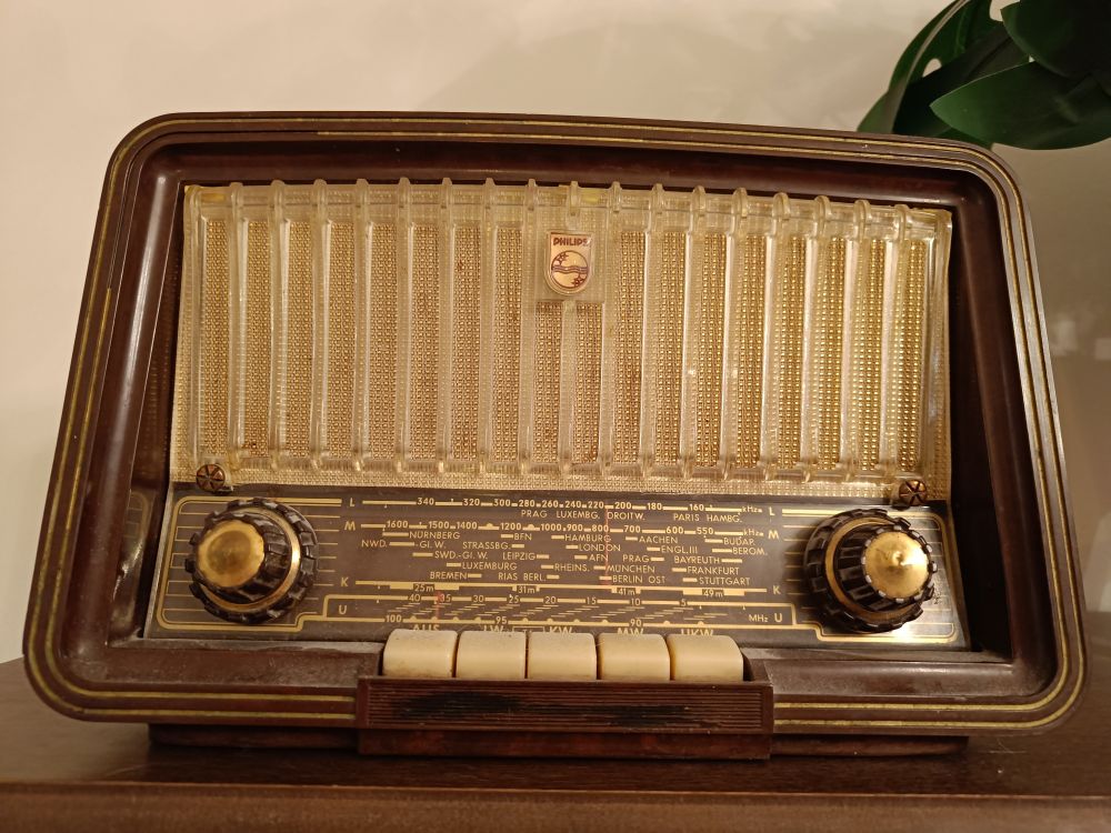 Vanha ruskea Philips-merkkinen putkiradio ruskean kaapin päällä, vieressä näkyy muovikukan lehti. Radion taajusvalitsin on kohdassa "BERLIN OST".