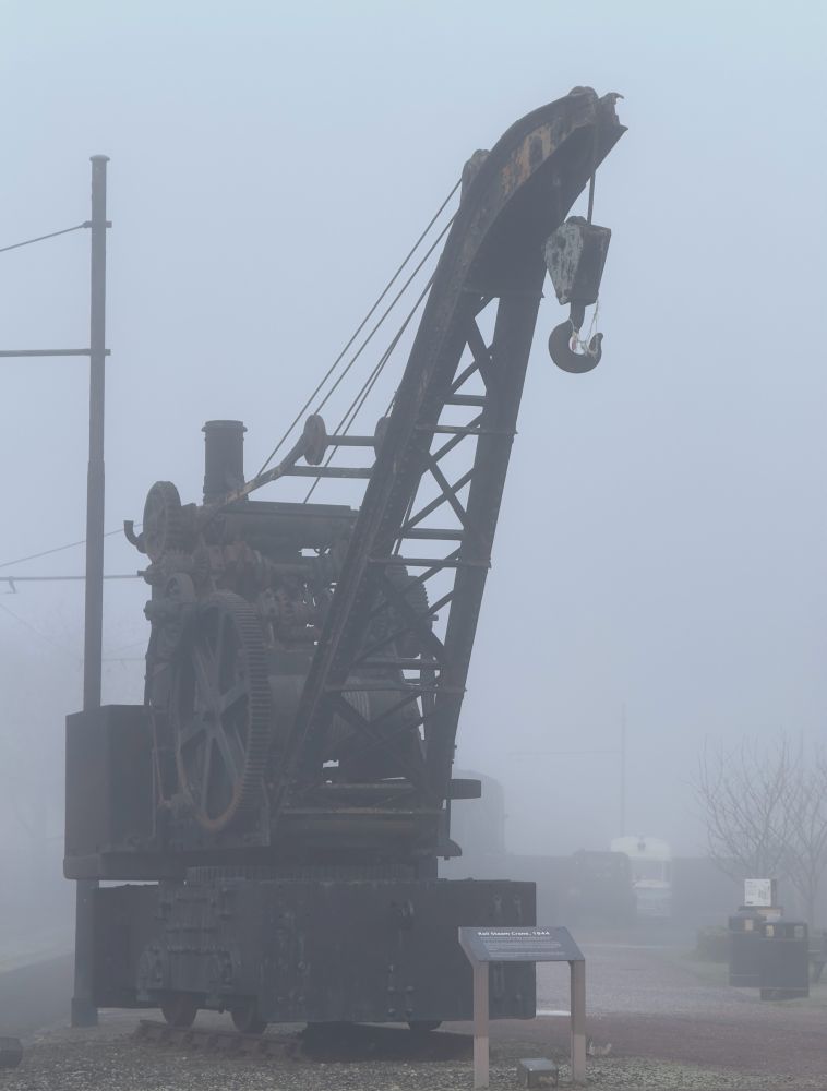 An old steam crane with hook hanging from its raised latticed steel jib, almost silhouetted against the misty background.