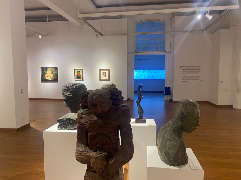 A few inside a white world, modern gallery space with paintings and text on the walls. In the centre foreground a series of sculptural busts stand on white rectangular plinths.
