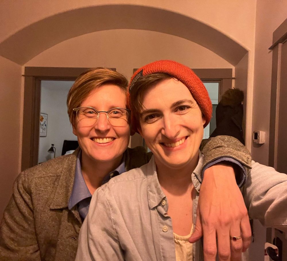 Two people smiling, one with glasses and dark blonde hair and grey jacket, and the other with dark hair leaking out from an orange beanie and light blue button-up shirt. The first person has her arm around the second, who is non-binary. Warm light. Indoors setting. 