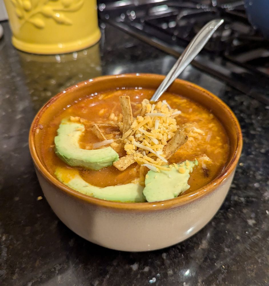 A tan bowl with a brown rim sits on a black counter with a spoon sticking out of it. Inside the bowl is a reddish-brown liquid with avocado slices, crispy tortilla strips, and white and yellow shredded cheese in it. 