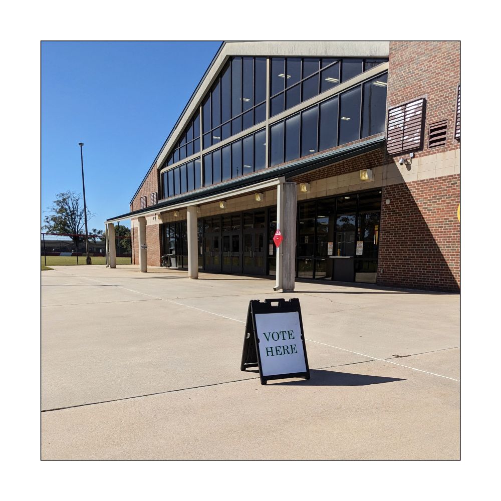 Small folding sign says: VOTE HERE. Sign sits in empty area in front of a large school building.