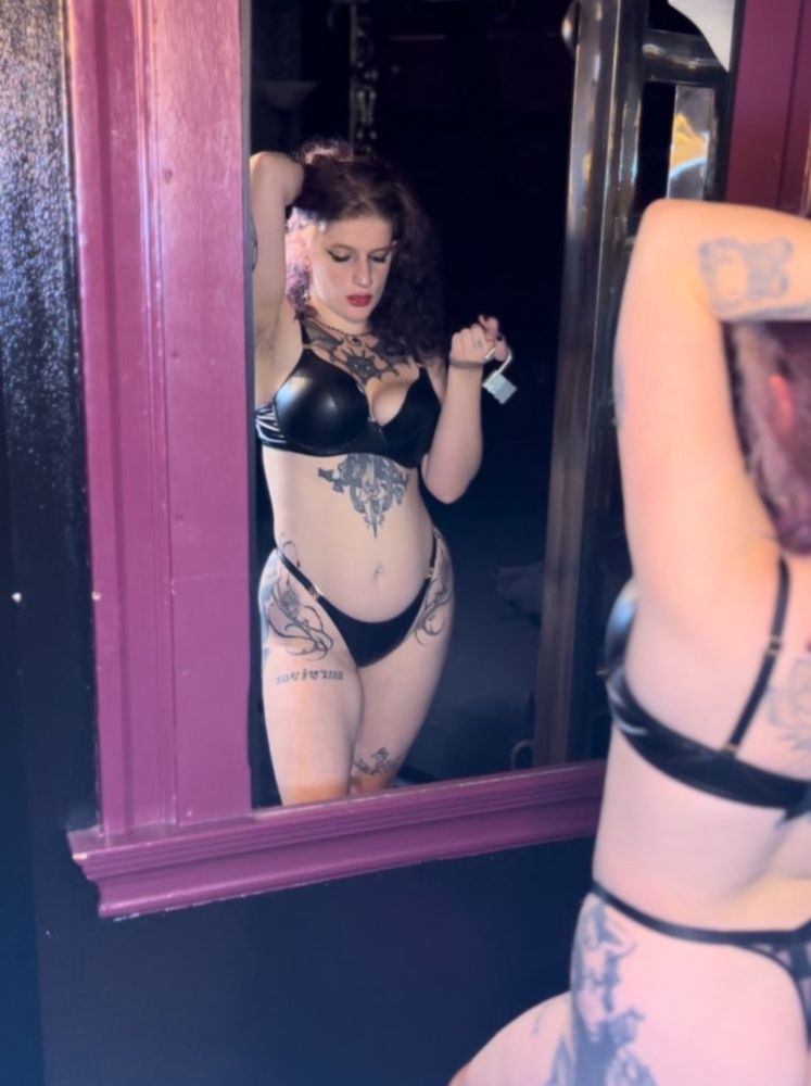Mxtress Lilin poses in front of a mirror in their old lair, holding up a padlock that will only be used to trap you further under their endless control. You are helpless against a goddess like them. 