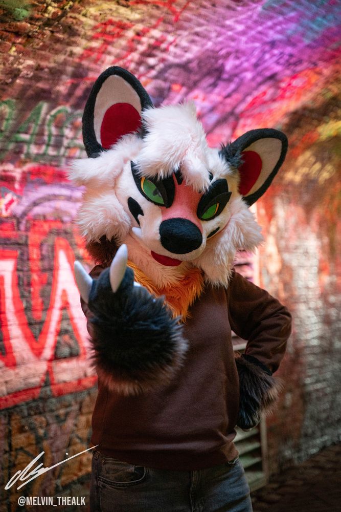 A marten fursuiter stands in a neon-lit tunnel covered in graffiti, looking at the camera and beckoning the viewer closer with their paw out.