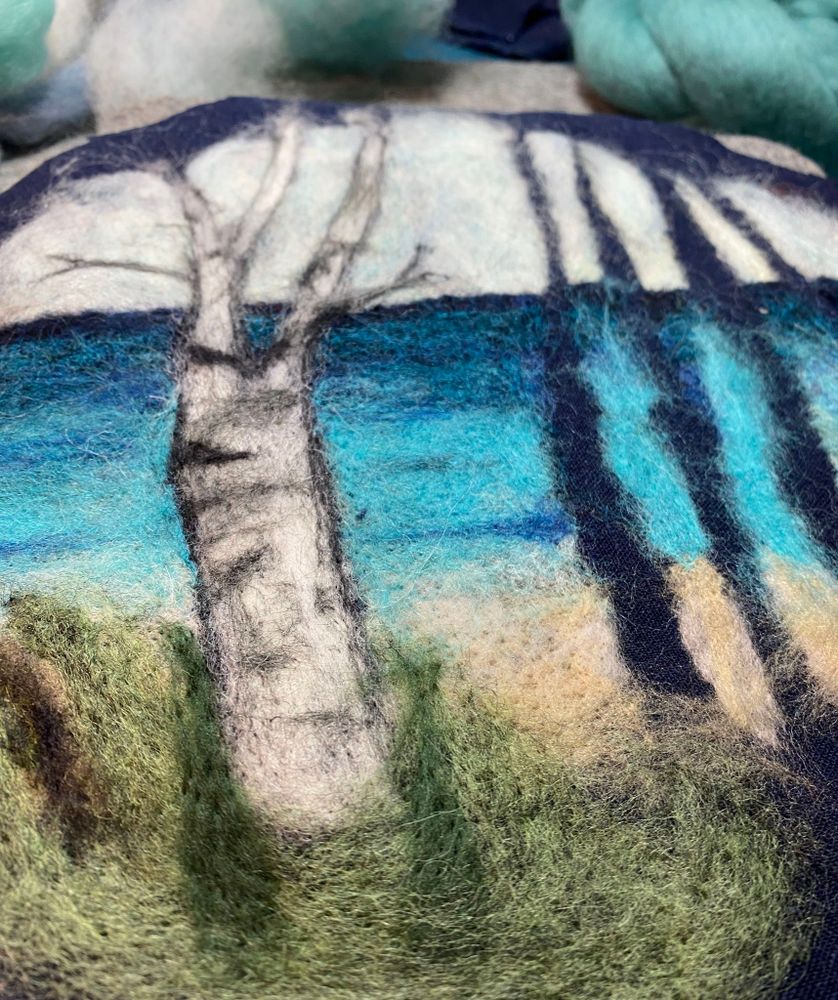 Needle felted wool landscape artwork on linen showing a birch tree with white bark in the foreground, set against layers of blue water or sky and green grassy ground, with darker blue and gray tones in the background