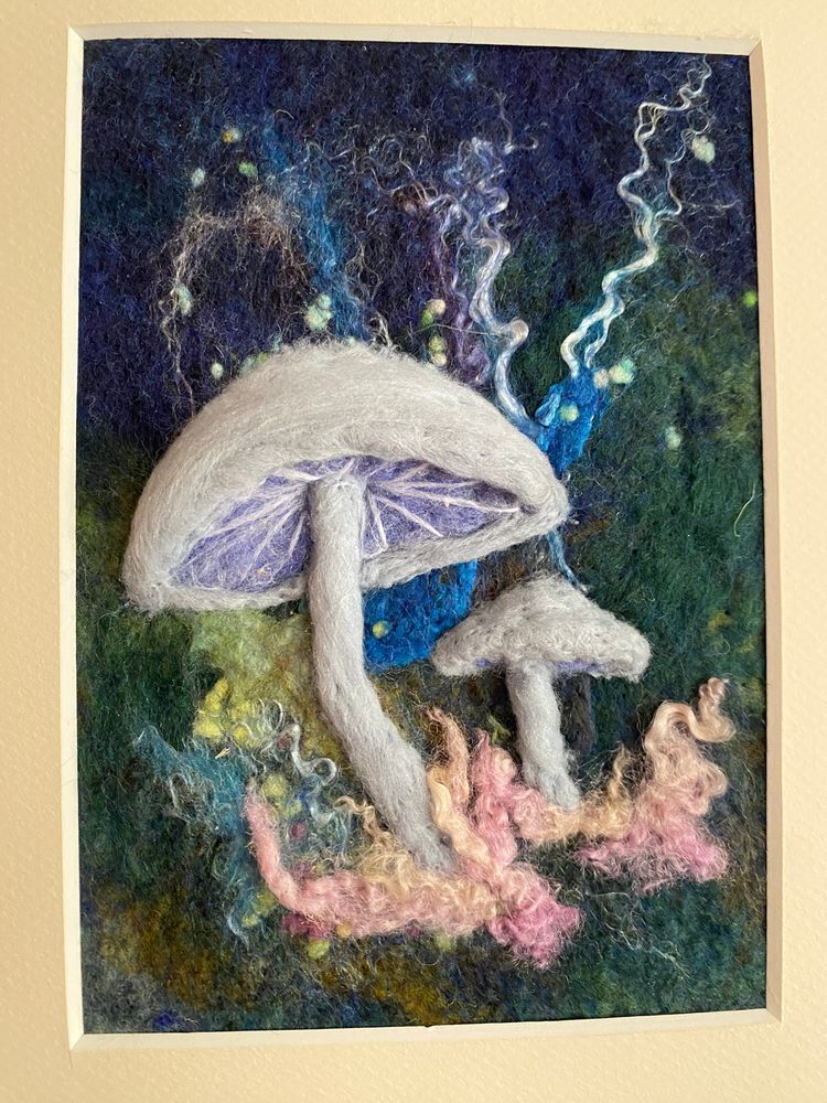 Needle felted artwork featuring two large white mushrooms with detailed gills on stems, surrounded by moss, lichen, and curly pink fiber at the base. The background is dark blue-green with white twisted branches and small green seed pods scattered throughout, creating a mystical forest floor scene.
