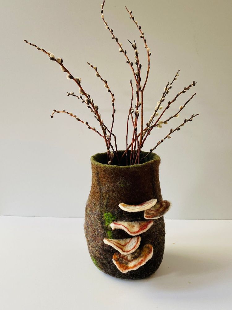 A brown wool vase, with 5 bracket mushrooms growing up the side. The vase is filled with twigs.