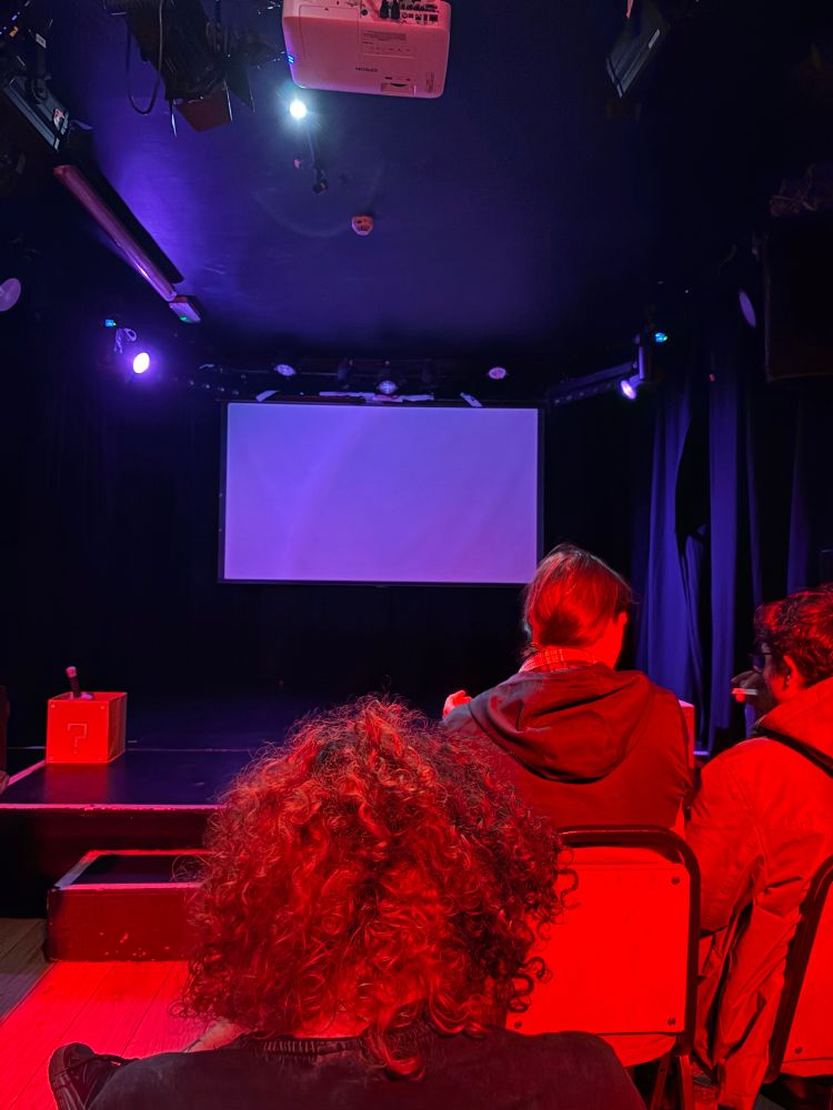 A crowd, bathed in red light, sit in front of a purple lit empty stage 