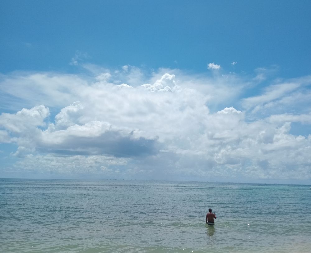 Solitary figure standing looking out to sea over the calm water with blue sky and clouds on the horizon.