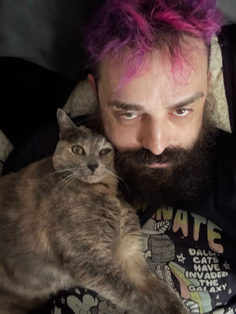 Cat laying on a bearded and magenta haired man’s chest looking at the ceiling picture taken from above so they are looking into the camera