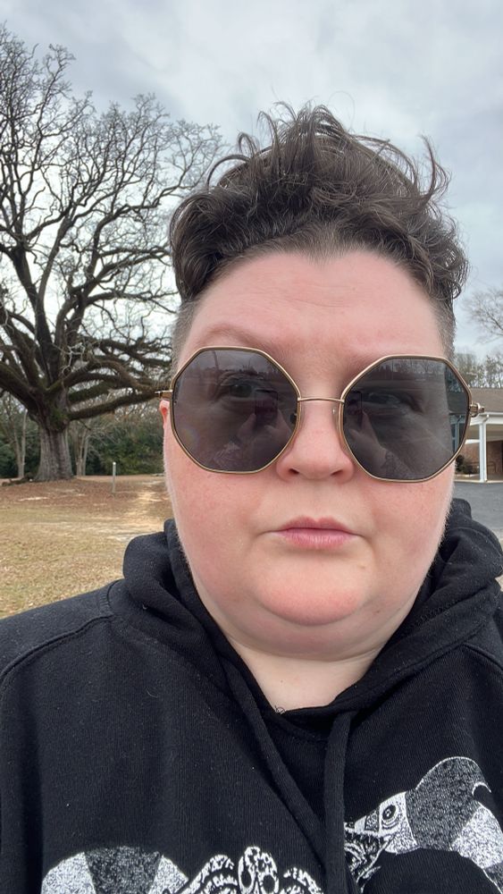 Pictured is a dark-haired, white non-binary person in a black hoodie sweatshirt and large sunglasses. They look unamused. Behind them, you can see a large, bare tree and some asphalt. The sky is overcast. 
