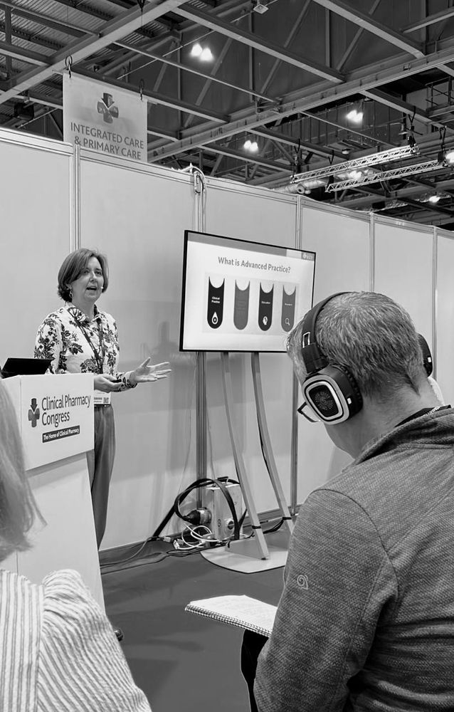 A black and white photograph of a person standing by a conference podium giving a talk. In the foreground is a man wearing headphones. A presentation slide showing the four pillars of advanced practice is visible in the background.