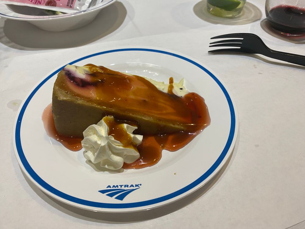 A slice of cheesecake is depicted including whipped cream and a berry syrup 