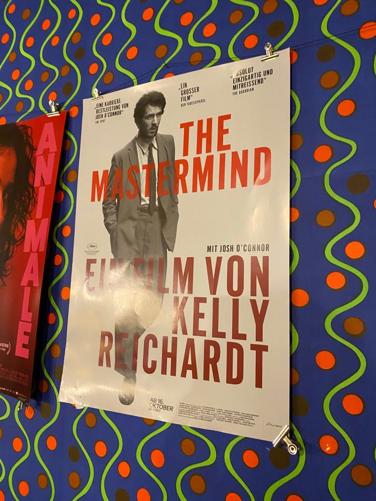 Das Filmposter von THE MASTERMIND an einer Wand mit grün-rot-blau gemusterter Tapete