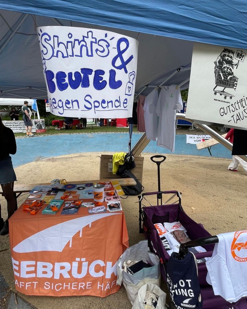 Foto eines Infostands mit Flyern, Stickern und Schlüsselbändern. Darüber hängt ein Schild, auf dem "Shirts & Beutel gegen Spende" steht. 