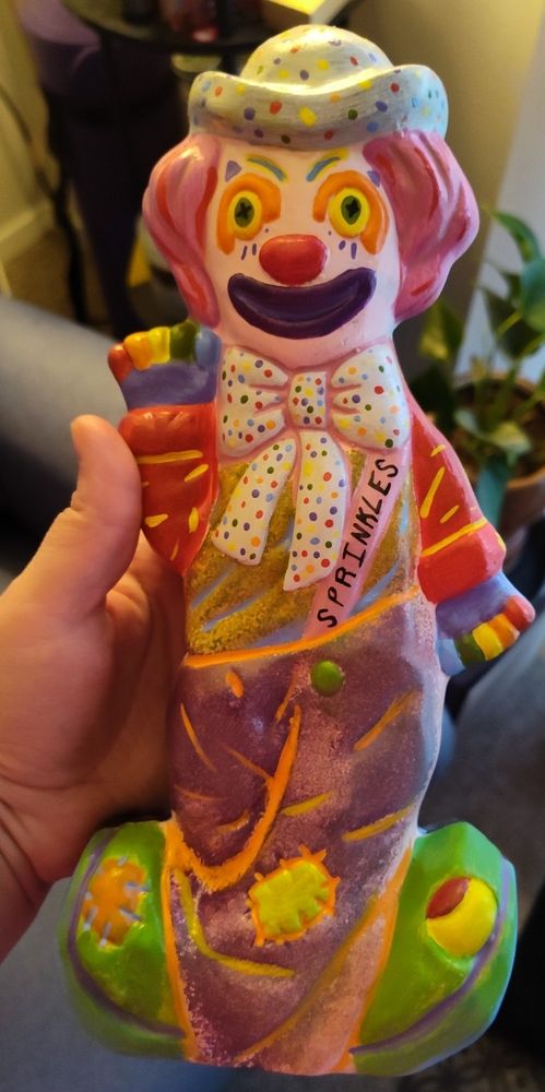 Painted ceramic smiling rainbow clown with rainbow polka dot hat, bow tie, and gloves. Multi colored shirt with red sleeves and yellow accents. Purple pants with yellow patch and orange accents. Green shoes with purple accents. Pink hair, rainbow clown makeup. The name 'Sprinkles' is written on pink sash.