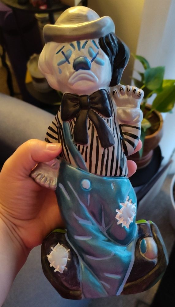 Blue, gray, silver, and black sad ceramic painted clown. Gray shirt with black horizontal stripes. Silver and gloves. Blue metallic pants with light blue accents. Black shoes with light blue and silver accents. Black bow tie. Sad blue clown makeup with tear running down cheek. Name 'Maury' on the fingers of the left hand.