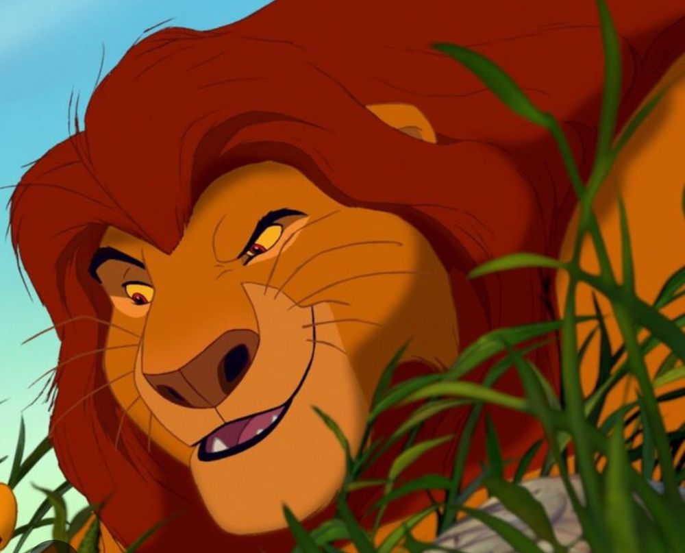 An orange lion with red mane and red eyes smiling coyly in grass 