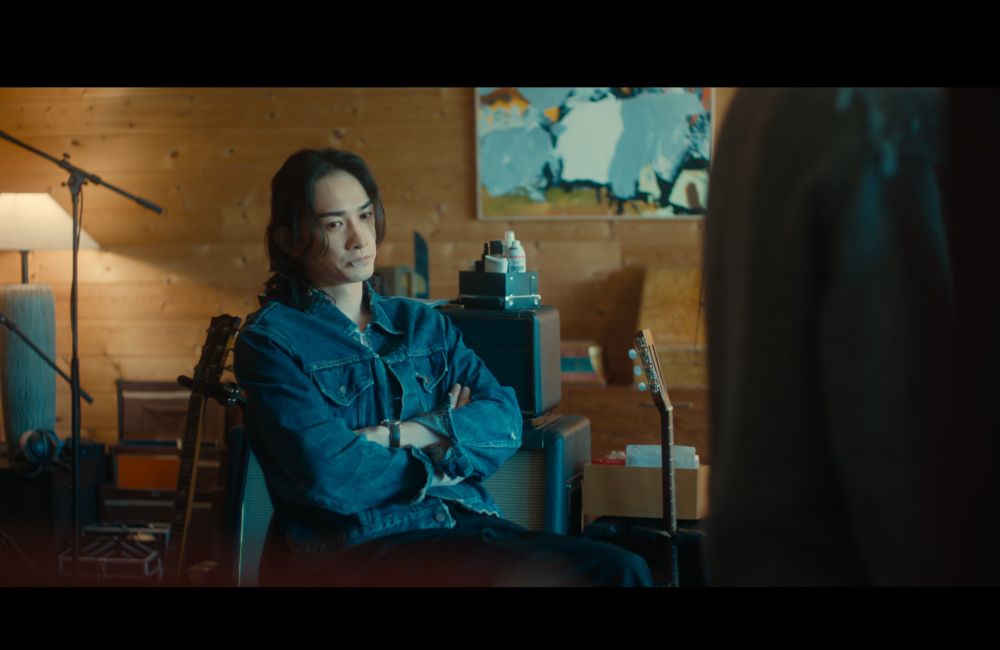 man in a blue denim shirt seated with arms crossed, in front of a wood-panelled wall. he's got a whiteish guitar pick in his mouth, and looks concerned