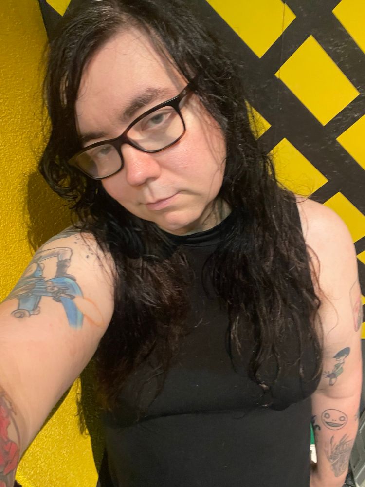 A girl with black hair and glasses wearing a tight sleeveless black top with tattoos on her arms 
