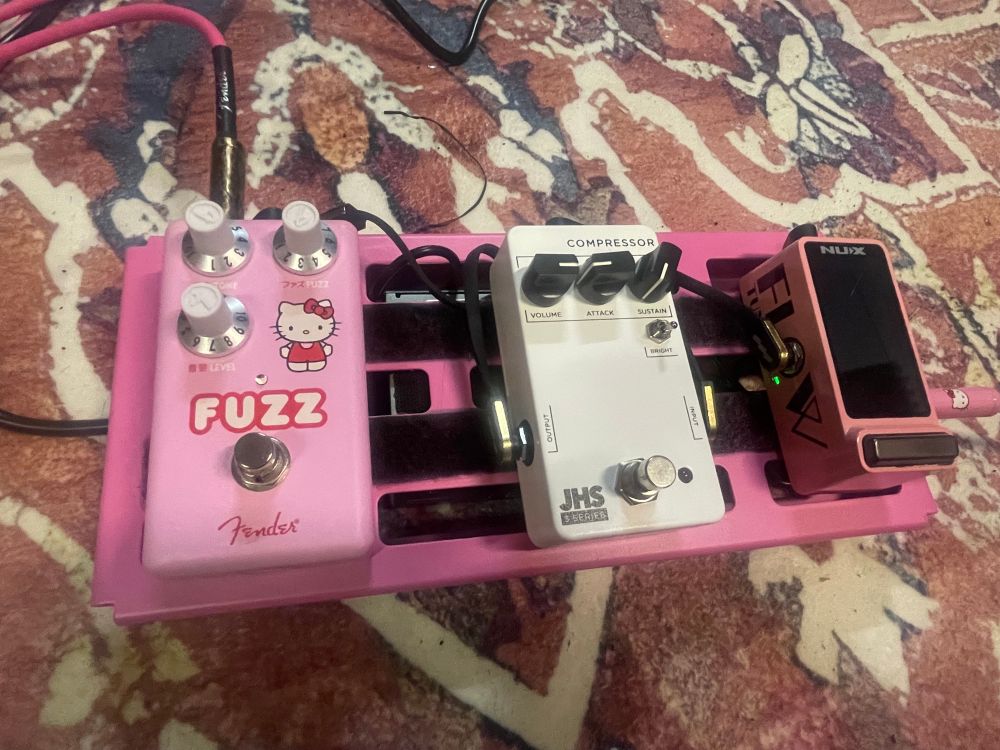 My Rockboard 2.0 pedal board, spray painted with Montana “Pink Cadillac” paint, and with black velcro strips. The pedals on it from left to right are: A pink Hello Kitty x Fender Fuzz Pedal, a JHS 3 Series Compressor, and a pink NU-X flow tuner. 