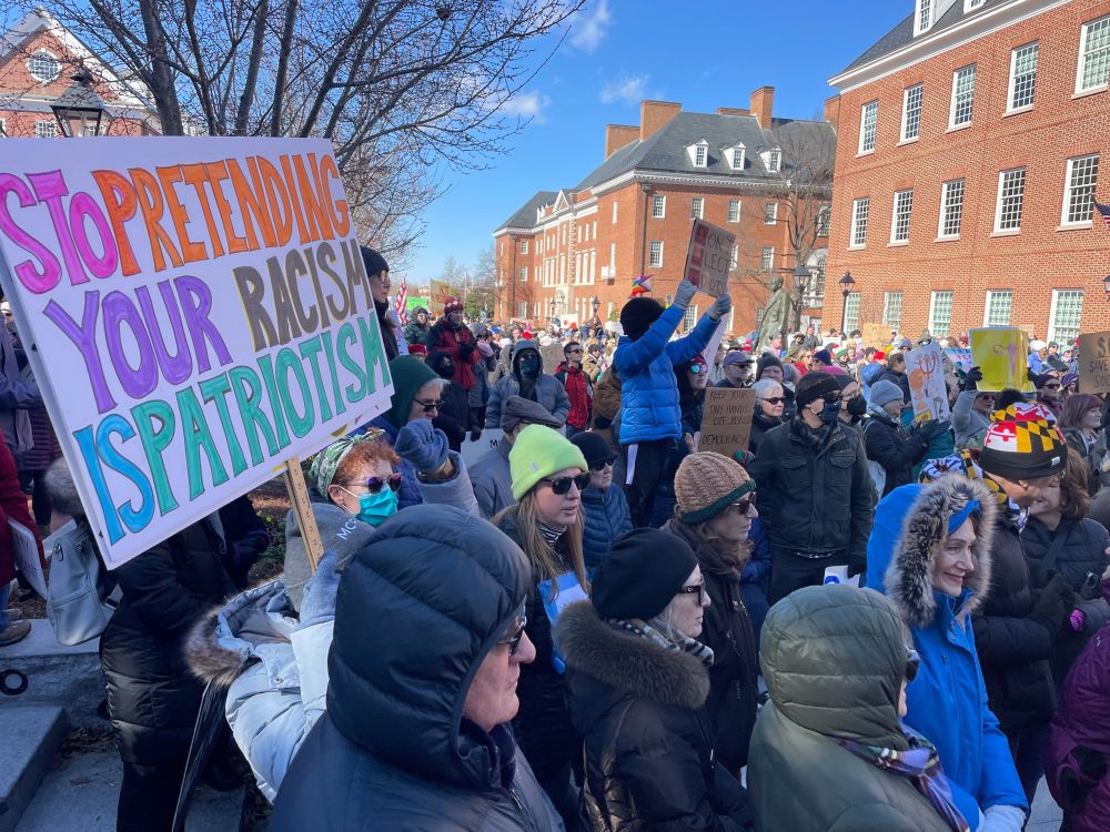 Crowd of people at a rally. Sign in the foreground reads: "Stop Pretending Your Racism Is Patriotism"