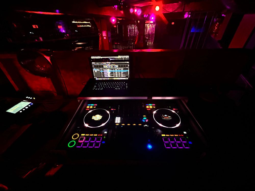 an empty bar floor with red and pink lighting. in the forefront is a dj controller seen only by its backlighting with a laptop seen only by the lit keys and screen. a song is qued up on the right jogwheel but not the left