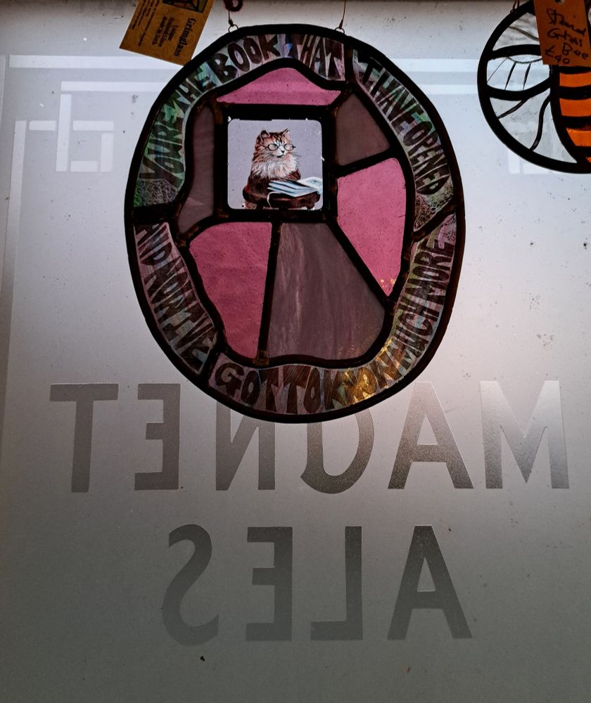 Stained glass panel, including a lyric from the Massive Attack song, 'Unfinished Sympathy', and a magic lantern slide of a cat with glasses on reading a book. The window it's hanging in has frosted glass, and the words 'Magnet Ales'.