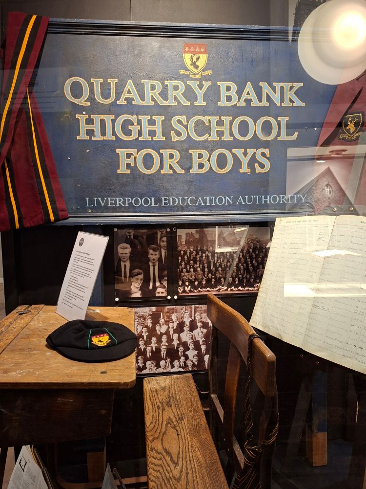 John Lennon's school desk, with a school cap from the period, school photos of Lennon, an attendance register featuring his name, and a sign reading 'Quarry Bank High School for Boys'.