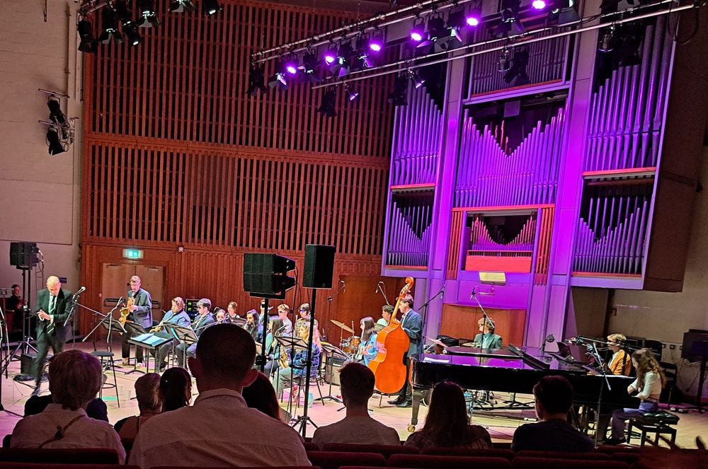 An 18-piece jazz band performing in the Jack Lyons concert hall at the University of York, with audience members in the foreground 