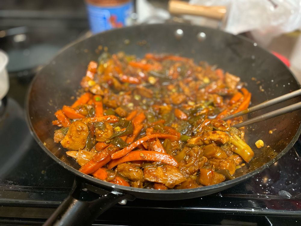 Large wok full of stir fry containing bite sized pieces of grilled chicken, carrot sticks, broccoli, corn, snap peas, and some other veggies