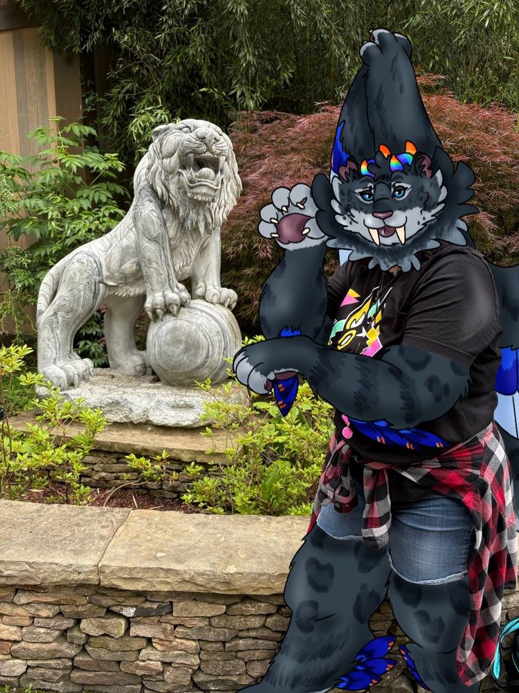 This is a drawover artwork of a picture I took of my partner at the zoo. Storm is a chubby snow leopard with rainbow horns and wings, featuring blue feathers with striped patterns like a  blue jay. She stands on the right side of the image in a “nya” pose with paws curled and one hand up by her face. She sticks her tongue out and smiles. They are wearing a black graphic tshirt with jean shorts and a black and red flannel tied around their waist. The background is mostly leaves, with a stone wall coming up to Storm’s thigh level. A stone statue of a tiger roaring is to the left of Storm.