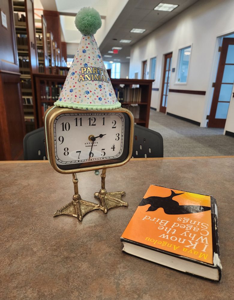 The duck feet clock, wearing a mini party hat that says "party animal", in a library reading I Know Why the Caged Bird Sings